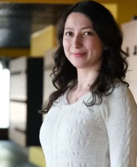 Uzun, koyu renkli ve dalgalı saçlı bir kadın, iç mekânda kameraya dönük şekilde hafifçe gülümsemektedir. Arka planda sarı, siyah ve nötr tonlarda mimari unsurlar bulanık biçimde görülmektedir.